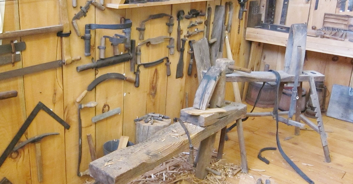 木工の手道具が壁一面に掛けられた作業場。手前に削りかけの木材と削り屑が散っている